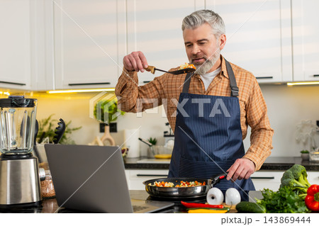 Middle-aged man cooks seasoned vegetables on electric stove watching laptop recipe for healthy diet Middle-aged man cooks seasoned vegetables on electric stove watching laptop recipe for healthy diet 143869444