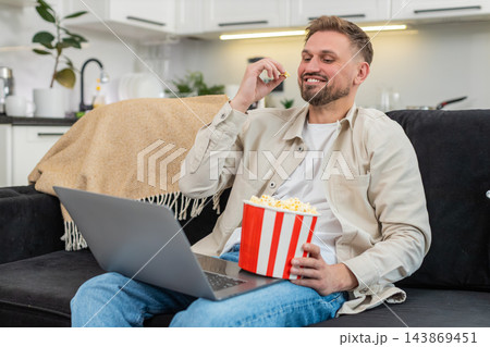 Young man watches comedy on laptop eats popcorn laughs out loud cheerful reaction happy movie time Young man watches comedy on laptop eats popcorn laughs out loud cheerful reaction happy movie time 143869451