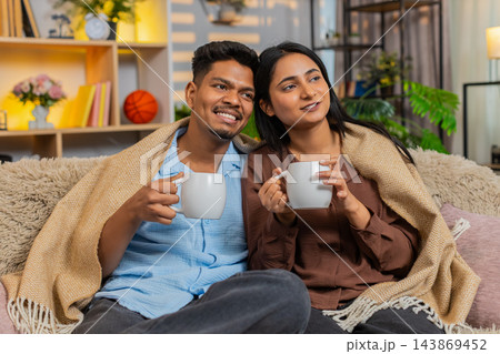 Indian couple man and woman sitting on home sofa with tea under blanket enjoying winter home comfort Indian couple man and woman sitting on home sofa with tea under blanket enjoying winter home comfort 143869452