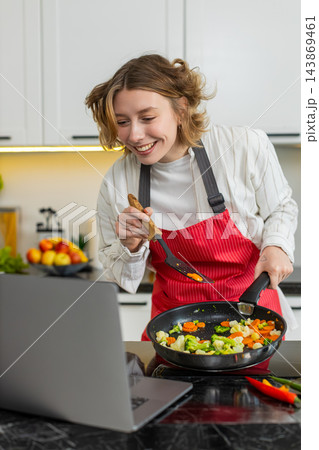Young woman cooking veggie stir fry while watching laptop recipe video for diet meal in home kitchen Young woman cooking veggie stir fry while watching laptop recipe video for diet meal in home kitchen 143869461