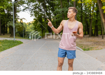Young man pointing sideways while holding orange smartphone and wearing a pink tank top in city park 143869484