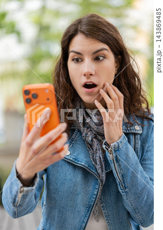 Young woman reacting with surprise while walking in city street looking at smartphone in amazement Young woman reacting with surprise while walking in city street looking at smartphone in amazement 143869485