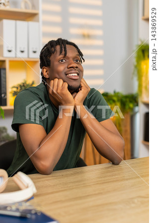 African American young man on home table resting on hands and daydreaming with dreamy smile indoors 143869529