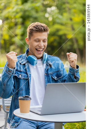 Young man celebrating work success and victory on laptop in city street with happy excited reaction 143869544