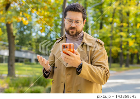 Disappointed man reading bad smartphone news with questioning hand gesture outdoors in autumn park 143869545