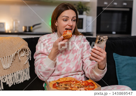 Adult woman enjoys pizza slice while texting on smartphone at night smiles cozy mood friendly chat Adult woman enjoys pizza slice while texting on smartphone at night smiles cozy mood friendly chat 143869547