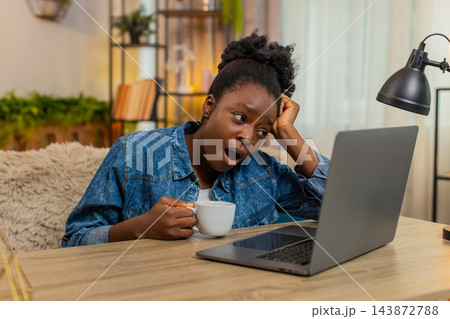 African American young woman drinks coffee tiredly before remote work on laptop sitting at home African American young woman drinks coffee tiredly before remote work on laptop sitting at home 143872788
