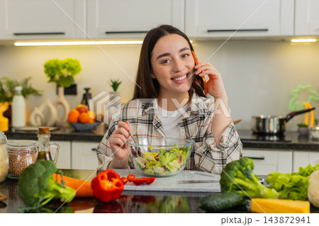 Young woman eating keto vegetable salad, talking on smartphone, sharing healthy recipe from kitchen Young woman eating keto vegetable salad, talking on smartphone, sharing healthy recipe from kitchen 143872941