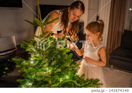 A woman and a girl arrange lights on a real Christmas tree in a living room. Decorating together prepares the home for Christmas and New Year. 143874149