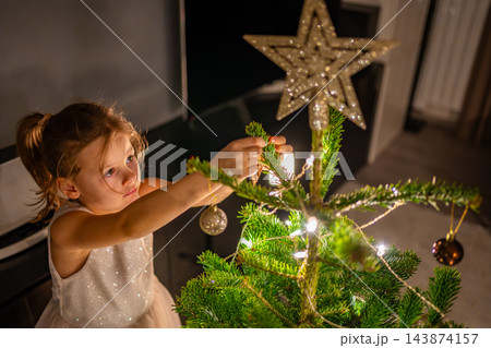 A girl hangs an ornament near the top of a glowing Christmas tree. Decorating the branches lets a child take part in New Year preparations. 143874157