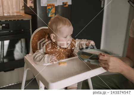 Caring mother hand feeding cute baby boy with spoon in high chair during mealtime at kitchen 143874547