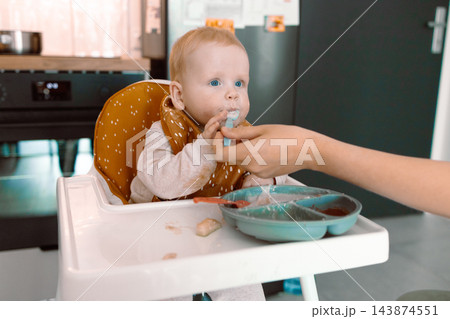 Caring mother hand feeding cute baby boy with spoon in high chair during mealtime at kitchen 143874551