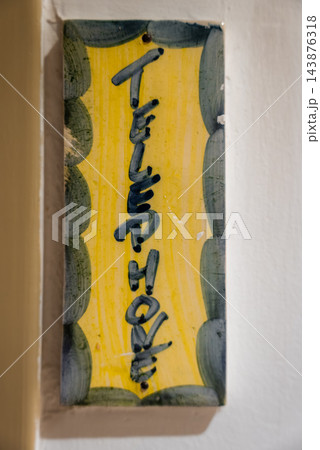 Vertical ceramic plaque reading Telephone hangs on a pale wall in Antibes, France. Soft indoor light reveals glaze, chips, and mottled green ovals framing yellow. 143876318
