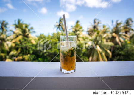 A glass containing a refreshing passion fruit cocktail with mint and ice sits on a white railing, framed by palm trees under a sunny sky in Dhigurah, Maldives. A glass containing a refreshing passion fruit cocktail with mint and ice sits on a white railing, framed by palm trees under a sunny sky in Dhigurah, Maldives. 143876329