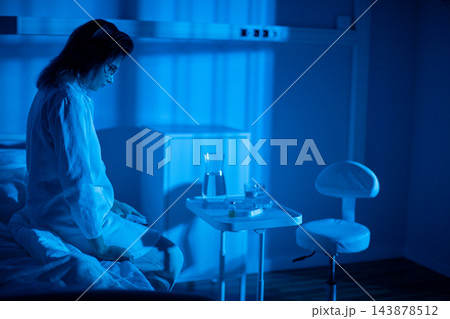 Patient sits on bed near window, pajamas wrinkled, lamp casting soft blue glow, hands folded while staring at distant lights, quiet moment of healing and reflection 143878512