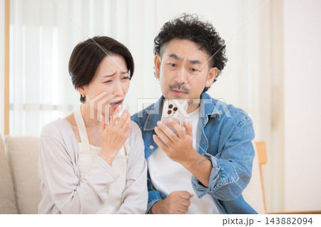 リビングでスマートフォンの画面を見ながら金利上昇や貯金、家計のニュースに驚く中年夫婦のライフスタイル 143882094