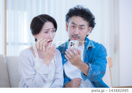 リビングでスマートフォンの画面を見ながら金利上昇や貯金、家計のニュースに驚く中年夫婦のライフスタイル 143882095