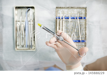 Gloved hand holds metal dental syringe above sterile instruments. Close up of anesthesia tool Gloved hand holds metal dental syringe above sterile instruments. Close up of anesthesia tool 143888229