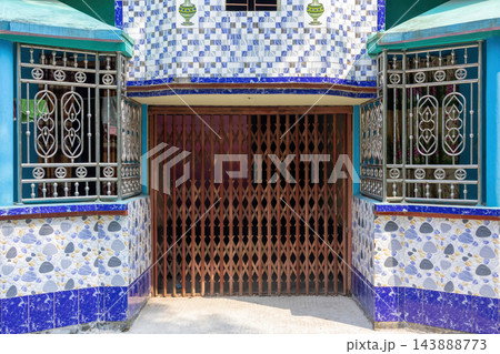 Front entrance of a residential building featuring a closed collapsible steel gate. Decorative stainless steel window grills and colorful ceramic wall tiles exterior. 143888773