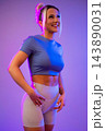Smiling athletic woman in workout clothes posing under neon purple and blue lighting 143890031