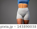 Fit woman in blue sports top and gray athletic shorts standing against gray background 143890038