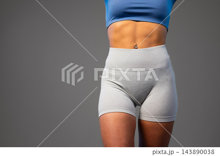 Fit woman in blue sports top and gray athletic shorts standing against gray background 143890038