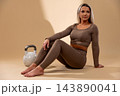 Athletic woman in brown workout outfit sitting with kettlebell on beige background 143890041