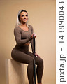 Fit young woman in brown workout outfit sitting on wooden box holding resistance band 143890043