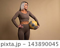 Fit woman in brown workout clothes holding a medicine ball in a studio setting 143890045