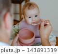Cute baby girl looking curiously into the camera while being fed by her father, highlighting early childhood nutrition, tender parent care, and love 143891339