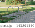 Outdoor stainless steel bike stands located in a quiet green park along a paved footpath under bright sunlight, representing cycling infrastructure 143891340
