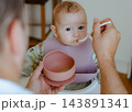 Sweet infant girl wearing a silicone bib taking a bite of solid food offered by dad, capturing authentic home lifestyle and parenting routine 143891341