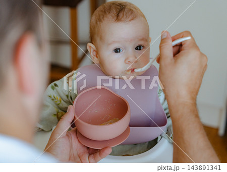 Sweet infant girl wearing a silicone bib taking a bite of solid food offered by dad, capturing authentic home lifestyle and parenting routine 143891341