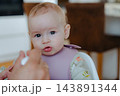 Cute baby girl with big brown eyes looking directly at camera while being fed by dad, capturing genuine facial expressions and child innocence 143891344