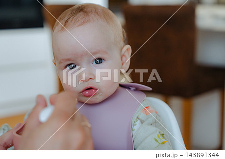 Cute baby girl with big brown eyes looking directly at camera while being fed by dad, capturing genuine facial expressions and child innocence 143891344