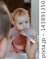 Adorable infant wearing a floral romper and bib looking attentively at father while eating solid food in a modern Scandinavian home interior 143891350