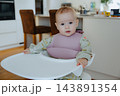 Adorable infant wearing a floral jumpsuit and silicone bib sitting calmly at a high chair table, capturing Scandinavian style nursery and home life 143891354