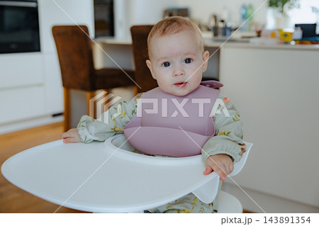 Adorable infant wearing a floral jumpsuit and silicone bib sitting calmly at a high chair table, capturing Scandinavian style nursery and home life Adorable infant wearing a floral jumpsuit and silicone bib sitting calmly at a high chair table, capturing Scandinavian style nursery and home life 143891354