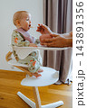 Elegant interior setup with an infant sitting in a white modern high chair while dad gives solid food using a spoon and silicone bowl 143891356