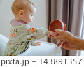 Curious little 7-8 month baby reaching out hands to touch a silicone bowl held by father during solid feeding session in a high chair, exploring food textures 143891357