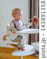 Full shot of an infant wearing a floral outfit and silicone bib enjoying solid food feeding with dad in a modern Nordic interior background 143891358