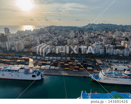 Durres Albania harbor features large passenger ferries moored beside container terminal. Dense urban buildings climb hillside above waterfront in warm evening light, with ships lined along coast. 143891428