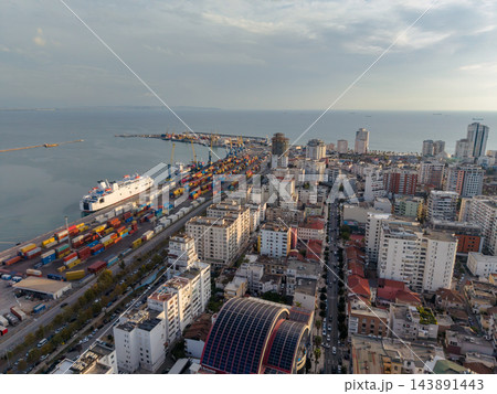 Durres Albania port sits beside dense urban waterfront, with cargo containers, towering cranes, and passenger vessel at berth. Calm Adriatic water and distant ships frame coastal city. Durres Albania port sits beside dense urban waterfront, with cargo containers, towering cranes, and passenger vessel at berth. Calm Adriatic water and distant ships frame coastal city. 143891443