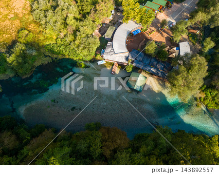 Blue Eye Albania natural spring winds through dense woodland, its brilliant blue pools bordering pebbled riverbed. Overhead perspective includes visitor buildings beside this striking scenic waterway. 143892557