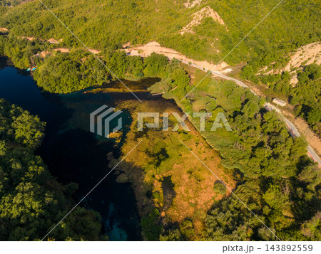 Blue Eye Albania spring lies amid dense green woodland, its deep blue water edged by golden vegetation. High aerial perspective shows surrounding hills, curving road and small riverside structure. Blue Eye Albania spring lies amid dense green woodland, its deep blue water edged by golden vegetation. High aerial perspective shows surrounding hills, curving road and small riverside structure. 143892559