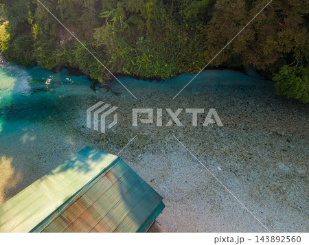 Blue Eye Albania spring reveals clear turquoise water over broad pebble bed. Dense woodland shades bank, while small wooden platform occupies foreground beside this vivid natural landmark. 143892560