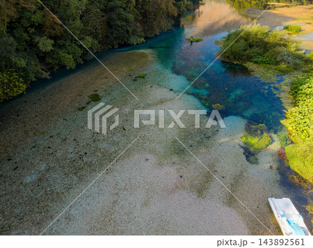 Blue Eye Albanias turquoise spring winds between dense woodland and sunlit vegetation. Crystal clear water reveals broad gravel bed, while small white boat rests beside natural river. Blue Eye Albanias turquoise spring winds between dense woodland and sunlit vegetation. Crystal clear water reveals broad gravel bed, while small white boat rests beside natural river. 143892561