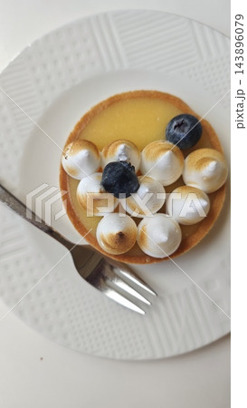 Round fruit tart with glossy yellow filling, blueberries, toasted meringue kisses, and fork on a white textured plate viewed from above, with circular crust and sweet baked pastry Round fruit tart with glossy yellow filling, blueberries, toasted meringue kisses, and fork on a white textured plate viewed from above, with circular crust and sweet baked pastry 143896079