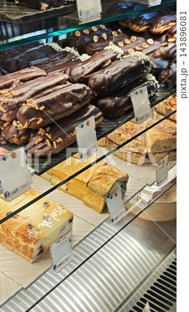 Assorted chocolate coated pastries and sliced cakes arranged in a glass bakery display with decorative topping details, labels, and layered desserts on illuminated shelves 143896081