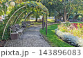 Curving brick walkway with benches beneath leafy trees, arched garden lights, green lawn, and colorful flower beds in a tranquil landscaped public park setting outdoors 143896083